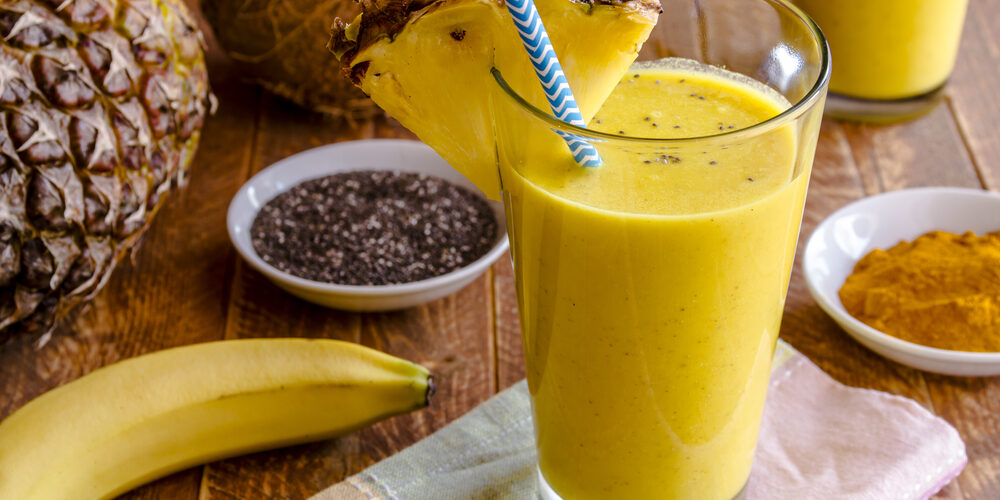 Close up fresh blended fruit smoothies made with pineapple, banana, coconut, turmeric and chia seeds surrounded by raw ingredients in drinking glass with pineapple slice garnish and blue striped straw